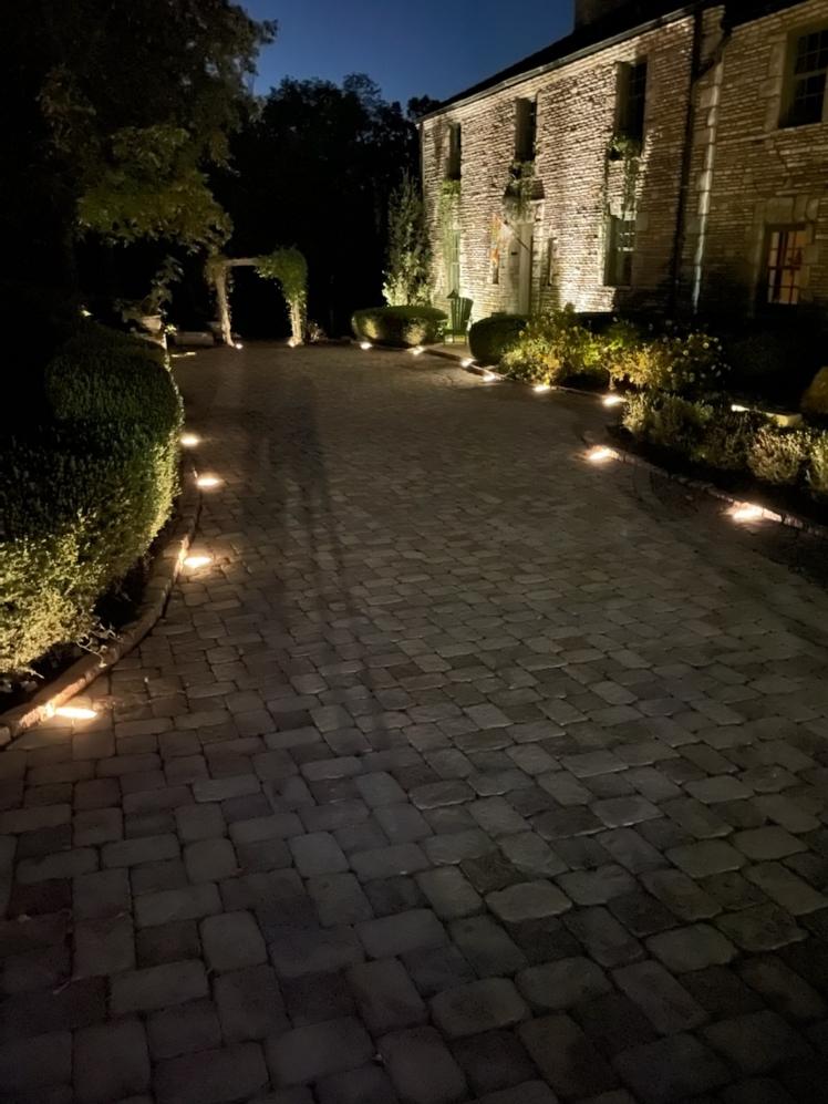 A paver drive at night, lit by low path lights, beside a stone house