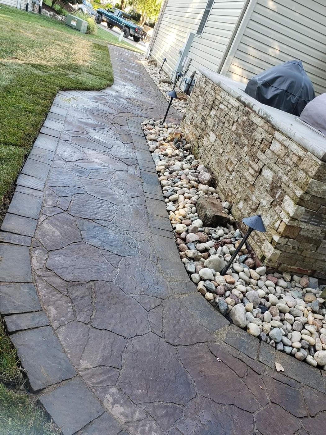 A flagstone-pattern paver walkway along a house with a river-rock bed and path lights